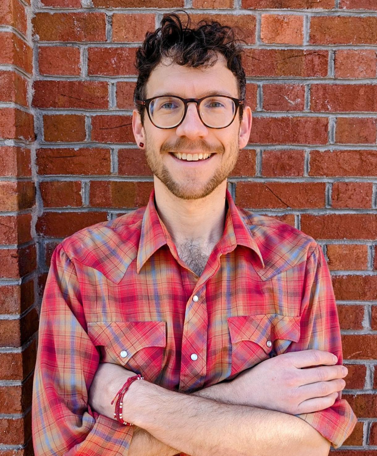 A student with arms crossed in a red plaid shirt and black glasses